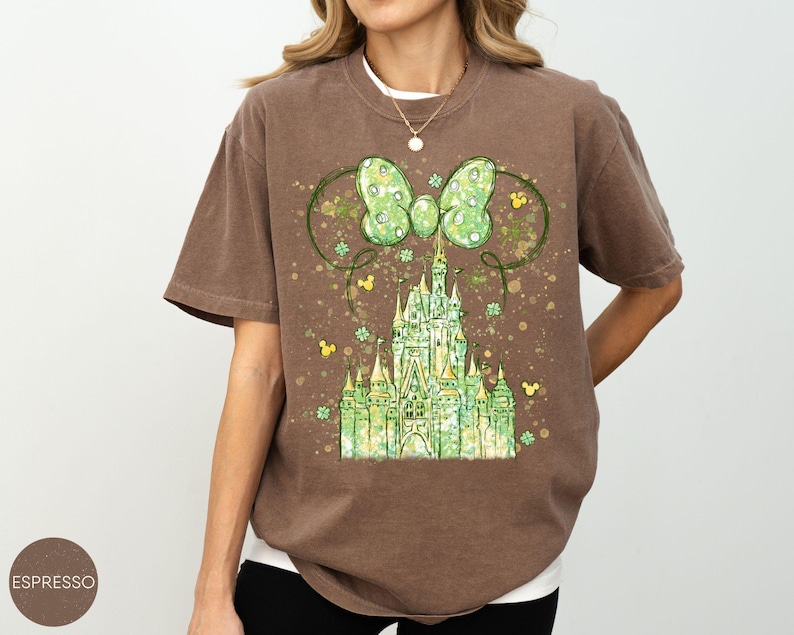 K&ouml;nnte beinhalten: Espresso-farbenes T-Shirt mit einem gr&uuml;nen Schloss-Design, Schleife und Mickey Mouse-Symbolen. Die Grafik verwendet Gr&uuml;n- und Gelbt&ouml;ne mit einem Vintage-Look. Das Shirt hat kurze &Auml;rmel und eine lockere Passform.