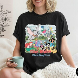 May include: A black t-shirt featuring a colorful graphic print of various Disney characters including Dumbo, Ariel, Cinderella, Mickey Mouse, and Goofy in front of a castle. The text Magic Kingdom and Walt Disney World is printed below the characters.
