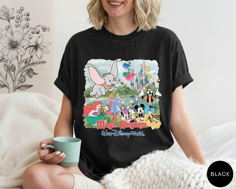 May include: A black t-shirt featuring a colorful graphic print of various Disney characters including Dumbo, Ariel, Cinderella, Mickey Mouse, and Goofy in front of a castle. The text Magic Kingdom and Walt Disney World is printed below the characters.