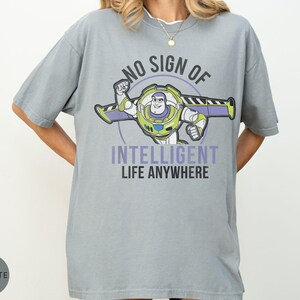 Puede incluir: Una camiseta gris con un dise&ntilde;o de Buzz Lightyear y el texto "NO SIGN OF INTELLIGENT LIFE ANYWHERE". El dise&ntilde;o presenta a Buzz en su traje espacial con detalles morados y verdes. La camiseta es de corte holgado.