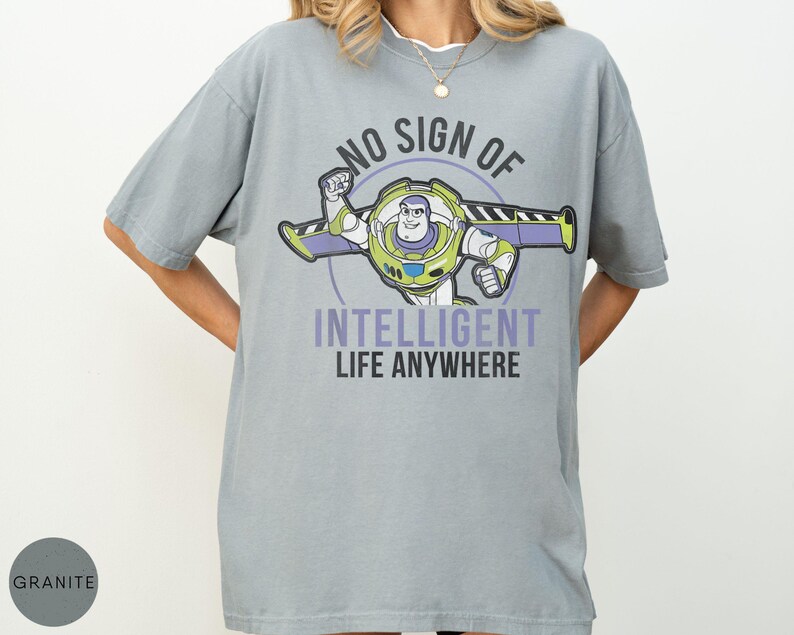 Puede incluir: Una camiseta gris con un dise&ntilde;o de Buzz Lightyear y el texto "NO SIGN OF INTELLIGENT LIFE ANYWHERE". El dise&ntilde;o presenta a Buzz en su traje espacial con detalles morados y verdes. La camiseta es de corte holgado.