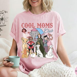 May include: Pink t-shirt with the text "COOL MOMS Club" in red and blue. The shirt features a colorful graphic of cartoon characters, including a lion cub, dogs, a cat, and women. The shirt is made of a soft material.
