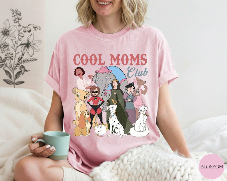 May include: Pink t-shirt with the text "COOL MOMS Club" in red and blue. The shirt features a colorful graphic of cartoon characters, including a lion cub, dogs, a cat, and women. The shirt is made of a soft material.