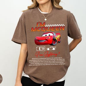 Puede incluir: Camiseta marr&oacute;n con el texto "I'M MCQUEEN" y un gr&aacute;fico de un coche de carreras rojo. La camiseta tambi&eacute;n incluye el texto "I am Lightning!" y una descripci&oacute;n del personaje. La camiseta es de corte holgado.