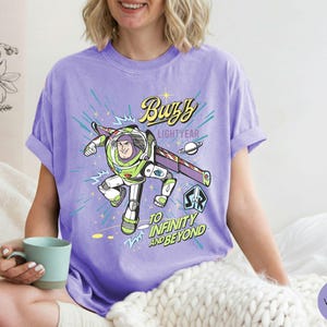 Peut inclure: T-shirt violet avec un motif de Buzz l'&Eacute;clair en vol. Le design comprend le texte "Buzz Lightyear" et "To Infinity and Beyond". Le t-shirt a des manches retrouss&eacute;es. L'image est d'une personne portant le t-shirt.