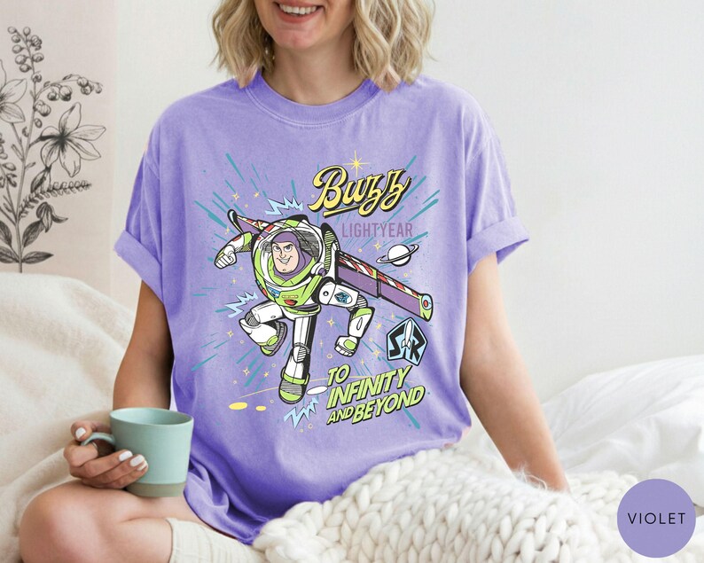 Peut inclure: T-shirt violet avec un motif de Buzz l'&Eacute;clair en vol. Le design comprend le texte "Buzz Lightyear" et "To Infinity and Beyond". Le t-shirt a des manches retrouss&eacute;es. L'image est d'une personne portant le t-shirt.