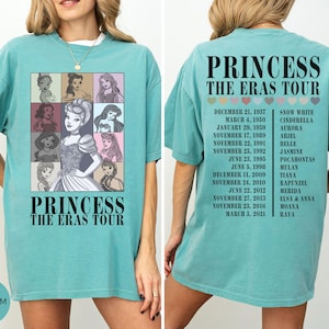 May include: Seafoam green t-shirt featuring a graphic of Disney princesses in a grid, with the text "PRINCESS THE ERAS TOUR." The back of the shirt lists tour dates and princess names. A casual, oversized fit.