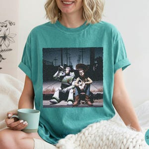 May include: A seafoam green short-sleeve t-shirt featuring a graphic print of two animated characters sitting on a curb at night. One character is a space ranger in a white and green suit, and the other is a cowboy wearing a brown hat and vest.