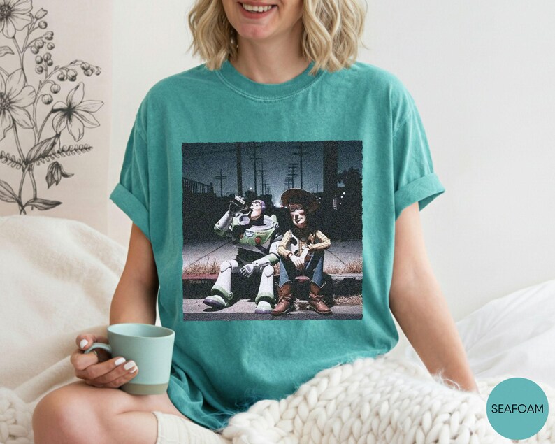May include: A seafoam green short-sleeve t-shirt featuring a graphic print of two animated characters sitting on a curb at night. One character is a space ranger in a white and green suit, and the other is a cowboy wearing a brown hat and vest.