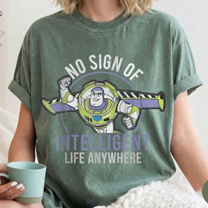 Puede incluir: Camiseta verde musgo con un gr&aacute;fico de Buzz Lightyear y el texto "NO SIGN OF INTELLIGENT LIFE ANYWHERE". El dise&ntilde;o incluye un contorno circular y el personaje en su pose caracter&iacute;stica.
