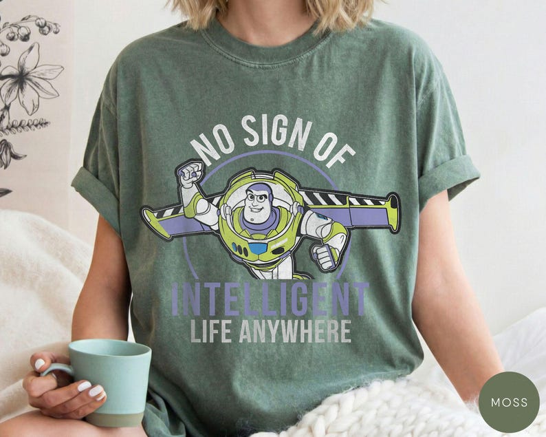 Puede incluir: Camiseta verde musgo con un gr&aacute;fico de Buzz Lightyear y el texto "NO SIGN OF INTELLIGENT LIFE ANYWHERE". El dise&ntilde;o incluye un contorno circular y el personaje en su pose caracter&iacute;stica.