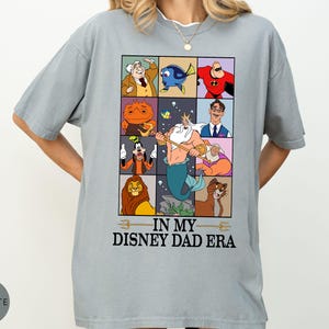 May include: A light gray t-shirt featuring a grid of Disney character illustrations. The shirt includes characters like Dory, Mr. Incredible, and Goofy. The text "IN MY DISNEY DAD ERA" is printed below the grid.