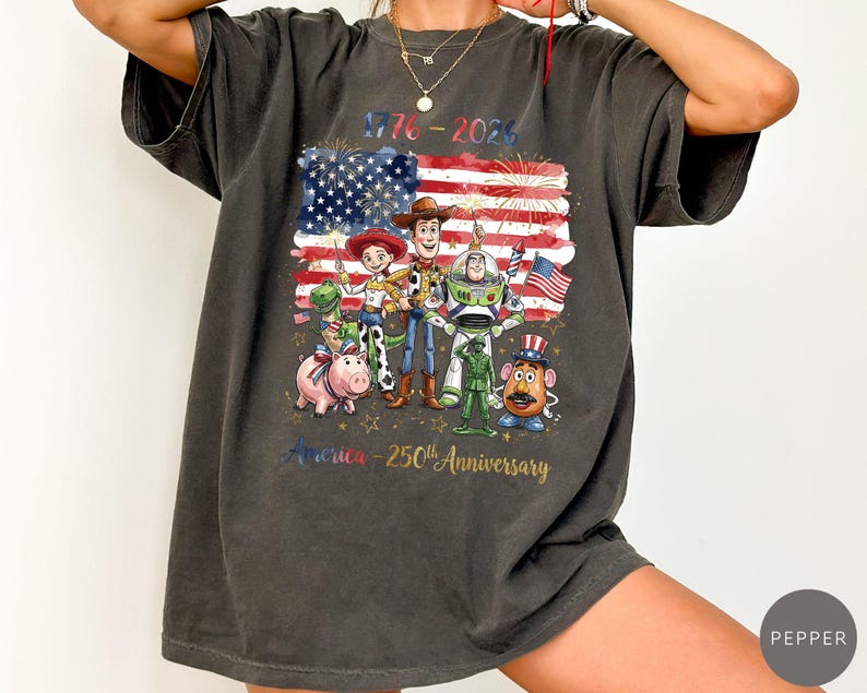 Pu&ograve; includere: T-shirt grigio scuro con una grafica della bandiera americana, fuochi d'artificio e personaggi dei cartoni animati. Il testo recita "1776-2026" e "America - 250th Anniversary".