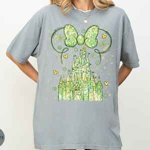 K&ouml;nnte beinhalten: Ein graues T-Shirt mit einem Aquarelldesign eines gr&uuml;nen Schlosses, einer Schleife und Mickey Mouse-Symbolen. Die Schleife hat ein Tupfenmuster. Das Schloss ist gr&uuml;n und gelb. Das Shirt hat eine lockere Passform.
