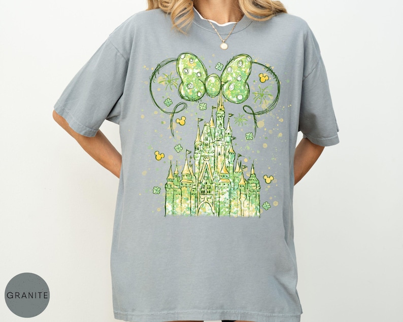 K&ouml;nnte beinhalten: Ein graues T-Shirt mit einem Aquarelldesign eines gr&uuml;nen Schlosses, einer Schleife und Mickey Mouse-Symbolen. Die Schleife hat ein Tupfenmuster. Das Schloss ist gr&uuml;n und gelb. Das Shirt hat eine lockere Passform.