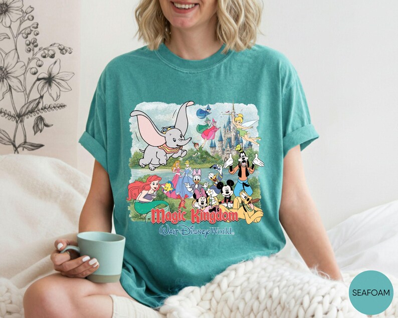 May include: A seafoam green short-sleeve t-shirt featuring a graphic print of Dumbo, Mickey Mouse, and other Disney characters in front of a castle. The text Magic Kingdom and Walt Disney World is printed in red and blue below the characters.