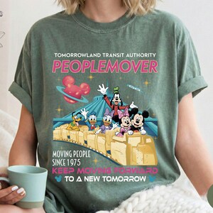 May include: A moss green t-shirt featuring a graphic of the Tomorrowland Transit Authority PeopleMover ride with Disney characters. The design includes the text "PEOPLEMOVER" and "MOVING PEOPLE SINCE 1975". The shirt has short sleeves and a relaxed fit.