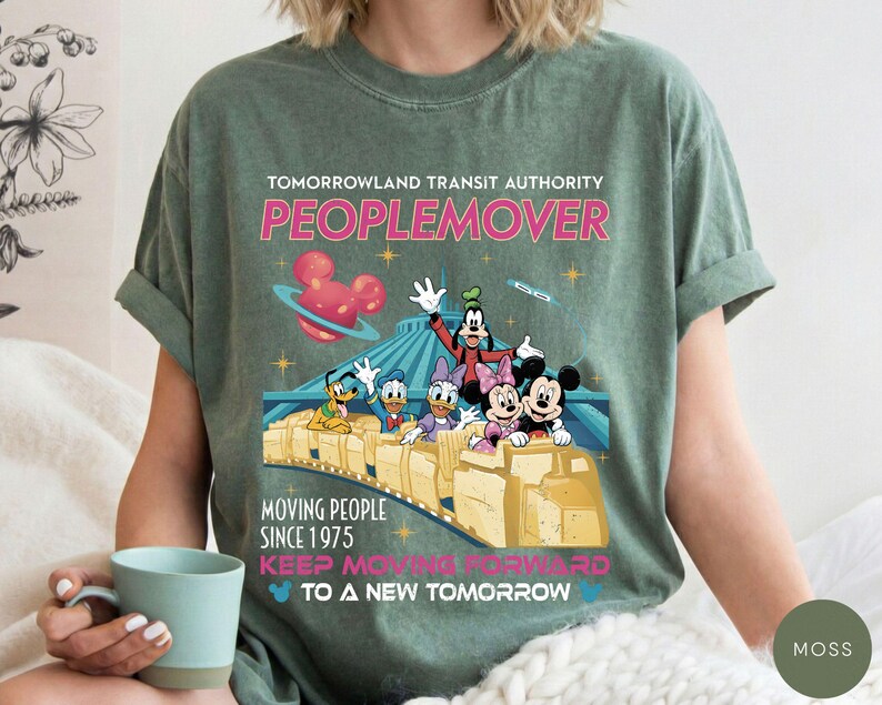 May include: A moss green t-shirt featuring a graphic of the Tomorrowland Transit Authority PeopleMover ride with Disney characters. The design includes the text "PEOPLEMOVER" and "MOVING PEOPLE SINCE 1975". The shirt has short sleeves and a relaxed fit.