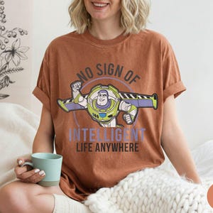 Puede incluir: Una camiseta de color &oacute;xido con un gr&aacute;fico de Buzz Lightyear y el texto "NO SIGN OF INTELLIGENT LIFE ANYWHERE". El dise&ntilde;o incluye a Buzz con su traje espacial con acentos verdes, morados y blancos. La camiseta tiene las mangas dobladas.