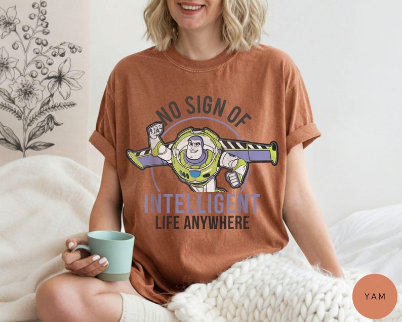 Puede incluir: Una camiseta de color &oacute;xido con un gr&aacute;fico de Buzz Lightyear y el texto "NO SIGN OF INTELLIGENT LIFE ANYWHERE". El dise&ntilde;o incluye a Buzz con su traje espacial con acentos verdes, morados y blancos. La camiseta tiene las mangas dobladas.
