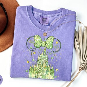 K&ouml;nnte beinhalten: Ein violettes T-Shirt mit einer gr&uuml;nen Schleife und einem Schlossdesign. Das Shirt hat einen Rundhalsausschnitt und kurze &Auml;rmel. Eine goldene Halskette mit Anh&auml;nger ist auf dem Shirt drapiert. Ein brauner Hut und getrocknete Palmbl&auml;tter sind im Hintergrund.