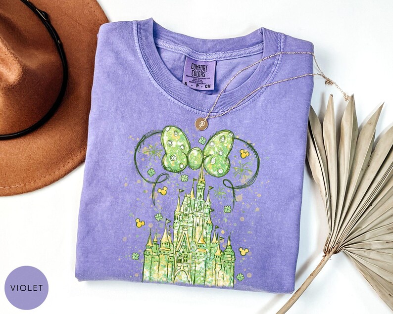 K&ouml;nnte beinhalten: Ein violettes T-Shirt mit einer gr&uuml;nen Schleife und einem Schlossdesign. Das Shirt hat einen Rundhalsausschnitt und kurze &Auml;rmel. Eine goldene Halskette mit Anh&auml;nger ist auf dem Shirt drapiert. Ein brauner Hut und getrocknete Palmbl&auml;tter sind im Hintergrund.