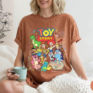 May include: A rust-coloured short-sleeve t-shirt featuring a colourful graphic print of Toy Story characters including Woody, Buzz Lightyear, Jessie, Rex, and others, with the Toy Story logo in red and yellow text centred above the character group.