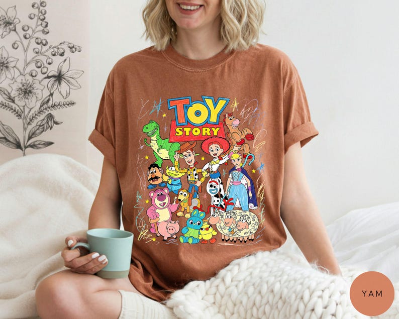 May include: A rust-coloured short-sleeve t-shirt featuring a colourful graphic print of Toy Story characters including Woody, Buzz Lightyear, Jessie, Rex, and others, with the Toy Story logo in red and yellow text centred above the character group.