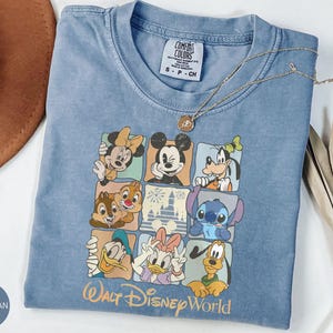 Może przedstawiać: Niebieska koszulka dżinsowa z kolorową grafiką postaci Disneya, w tym Myszki Miki i Myszki Minnie. Koszulka ma okrągły dekolt i kr&oacute;tkie rękawy. U dołu nadrukowano napis "Walt Disney World".