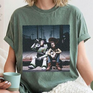 May include: A moss green short-sleeve t-shirt featuring a graphic print of Buzz Lightyear and Woody sitting on a curb at night. Buzz is drinking from a bottle while Woody sits beside him. The scene is set against a dark, urban street background.