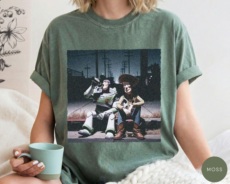 May include: A moss green short-sleeve t-shirt featuring a graphic print of Buzz Lightyear and Woody sitting on a curb at night. Buzz is drinking from a bottle while Woody sits beside him. The scene is set against a dark, urban street background.