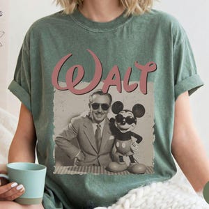 Puede incluir: Camiseta verde musgo con un gr&aacute;fico vintage de Walt Disney y Mickey Mouse. La palabra "Walt" est&aacute; escrita en rosa sobre la imagen. La camiseta tiene mangas cortas y cuello redondo.