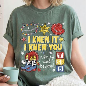May include: A moss green short-sleeve t-shirt featuring a graphic print with a sheriff badge, a red cowboy hat, a beaded bracelet, cowboy boots, and alphabet blocks. The text reads I KNEW IT I KNEW YOU infinity and beyond.