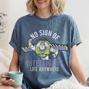 Puede incluir: Camiseta azul vaquero con un gr&aacute;fico de Buzz Lightyear y el texto "NO SIGN OF INTELLIGENT LIFE ANYWHERE". El dise&ntilde;o presenta a Buzz en su pose ic&oacute;nica, con detalles verdes, morados y blancos. La camiseta tiene las mangas dobladas.