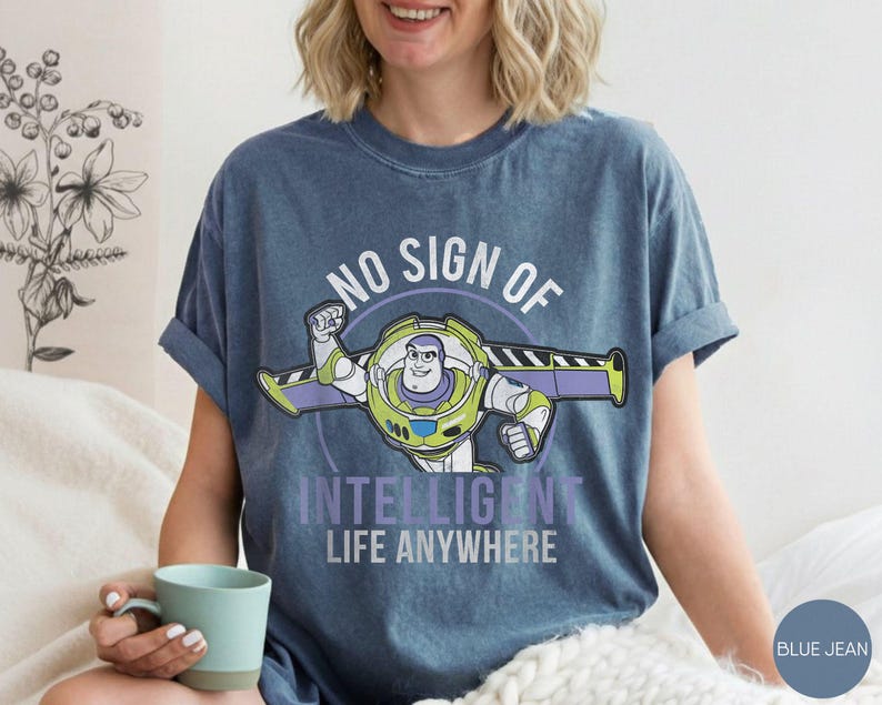 Puede incluir: Camiseta azul vaquero con un gr&aacute;fico de Buzz Lightyear y el texto "NO SIGN OF INTELLIGENT LIFE ANYWHERE". El dise&ntilde;o presenta a Buzz en su pose ic&oacute;nica, con detalles verdes, morados y blancos. La camiseta tiene las mangas dobladas.