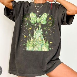 K&ouml;nnte beinhalten: Dunkelgraues T-Shirt mit einem gr&uuml;nen Schlossdesign, einer Schleife und Mickey Mouse Ohren. Das Design umfasst gr&uuml;ne Kleebl&auml;tter und gelbe Akzente. Das Shirt hat eine lockere Passform und kurze &Auml;rmel.