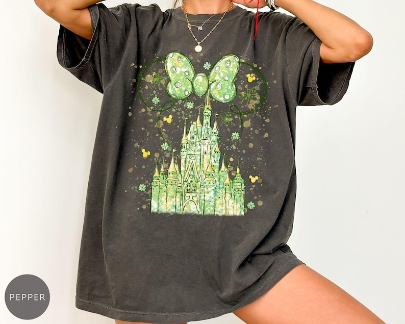 K&ouml;nnte beinhalten: Dunkelgraues T-Shirt mit einem gr&uuml;nen Schlossdesign, einer Schleife und Mickey Mouse Ohren. Das Design umfasst gr&uuml;ne Kleebl&auml;tter und gelbe Akzente. Das Shirt hat eine lockere Passform und kurze &Auml;rmel.