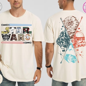May include: Ivory-colored t-shirt featuring the Star Wars logo with lightsabers and character illustrations. The back of the shirt displays a Death Star, Darth Vader, a Stormtrooper, and a TIE fighter in red, blue, and brown.