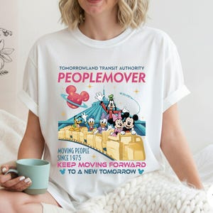 May include: White t-shirt featuring a colorful graphic of Disney characters on the Tomorrowland PeopleMover ride. The design includes the text "TOMORROWLAND TRANSIT AUTHORITY PEOPLEMOVER" and "MOVING PEOPLE SINCE 1975".