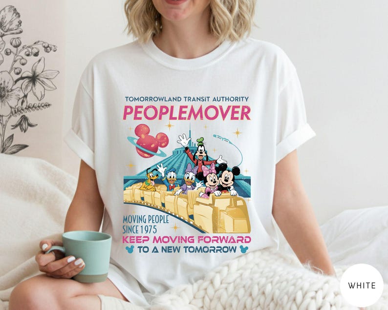 May include: White t-shirt featuring a colorful graphic of Disney characters on the Tomorrowland PeopleMover ride. The design includes the text "TOMORROWLAND TRANSIT AUTHORITY PEOPLEMOVER" and "MOVING PEOPLE SINCE 1975".