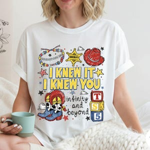 May include: A white t-shirt featuring a colorful graphic print with the text I KNEW IT I KNEW YOU infinity and beyond. The design includes illustrations of a beaded bracelet, a yellow sheriff star, a red cowboy hat, cowboy boots, and blocks.