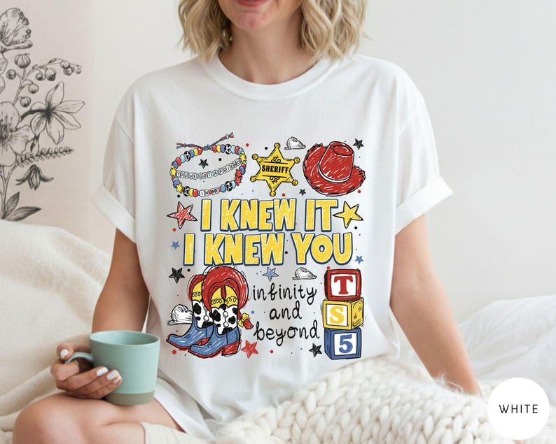 May include: A white t-shirt featuring a colorful graphic print with the text I KNEW IT I KNEW YOU infinity and beyond. The design includes illustrations of a beaded bracelet, a yellow sheriff star, a red cowboy hat, cowboy boots, and blocks.