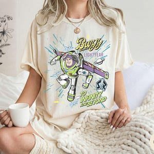 Peut inclure: T-shirt ivoire avec un motif de Buzz l'&Eacute;clair, le texte indique "Buzz Lightyear" et "To Infinity and Beyond". Le motif pr&eacute;sente une image de dessin anim&eacute; de Buzz l'&Eacute;clair dans sa combinaison spatiale, avec des accents bleus et verts.