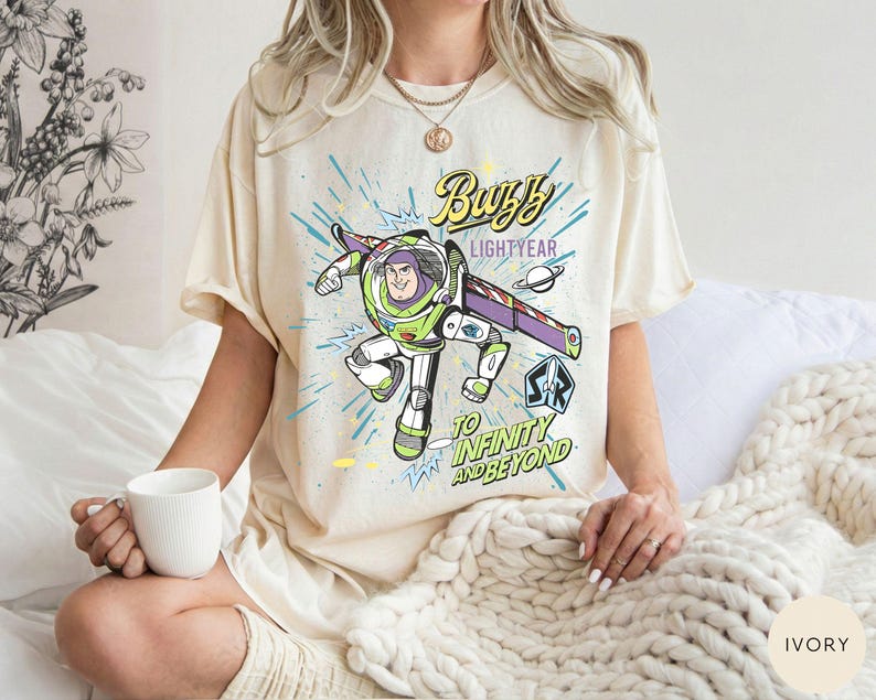 Peut inclure: T-shirt ivoire avec un motif de Buzz l'&Eacute;clair, le texte indique "Buzz Lightyear" et "To Infinity and Beyond". Le motif pr&eacute;sente une image de dessin anim&eacute; de Buzz l'&Eacute;clair dans sa combinaison spatiale, avec des accents bleus et verts.