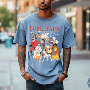 May include: Blue jean-colored t-shirt with the text "COOL DADS CLUB" in red and white. The shirt features a graphic of cartoon characters. The model is wearing the shirt outdoors.