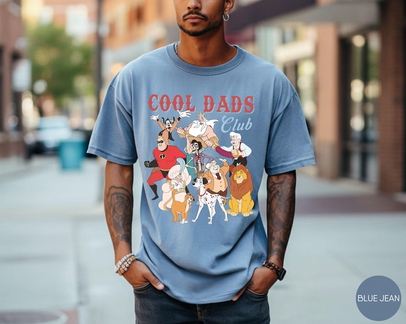 May include: Blue jean-colored t-shirt with the text "COOL DADS CLUB" in red and white. The shirt features a graphic of cartoon characters. The model is wearing the shirt outdoors.