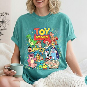 May include: A seafoam green short-sleeved t-shirt featuring a colourful graphic print of Toy Story characters including Woody, Buzz Lightyear, Jessie, Rex, and others, with the Toy Story logo at the top and scattered crayon-like star drawings.