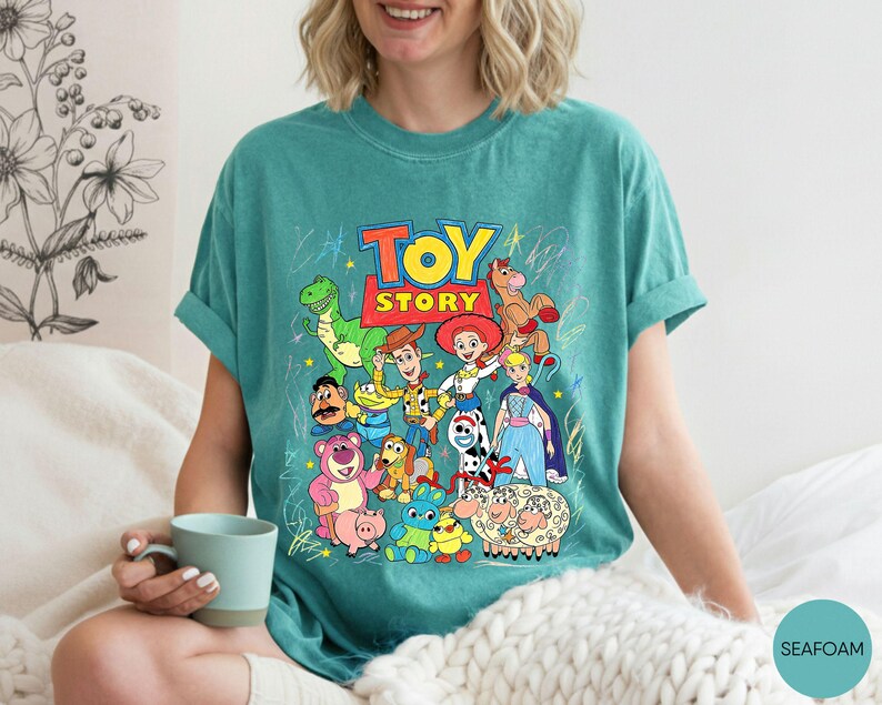 May include: A seafoam green short-sleeved t-shirt featuring a colourful graphic print of Toy Story characters including Woody, Buzz Lightyear, Jessie, Rex, and others, with the Toy Story logo at the top and scattered crayon-like star drawings.