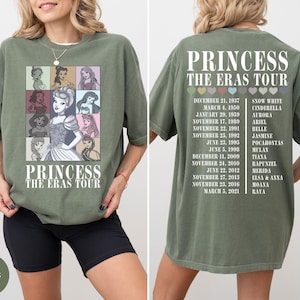 May include: Moss green t-shirt featuring a graphic of Disney princesses and the text "PRINCESS THE ERAS TOUR." The back of the shirt lists princess names and tour dates. The shirt is a comfortable, casual style.