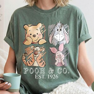 May include: Moss green t-shirt featuring four cartoon character portraits: Winnie the Pooh, Eeyore, Tigger, and Piglet. The words "POOH & CO. EST. 1926" are printed below the characters. The shirt has rolled-up sleeves.